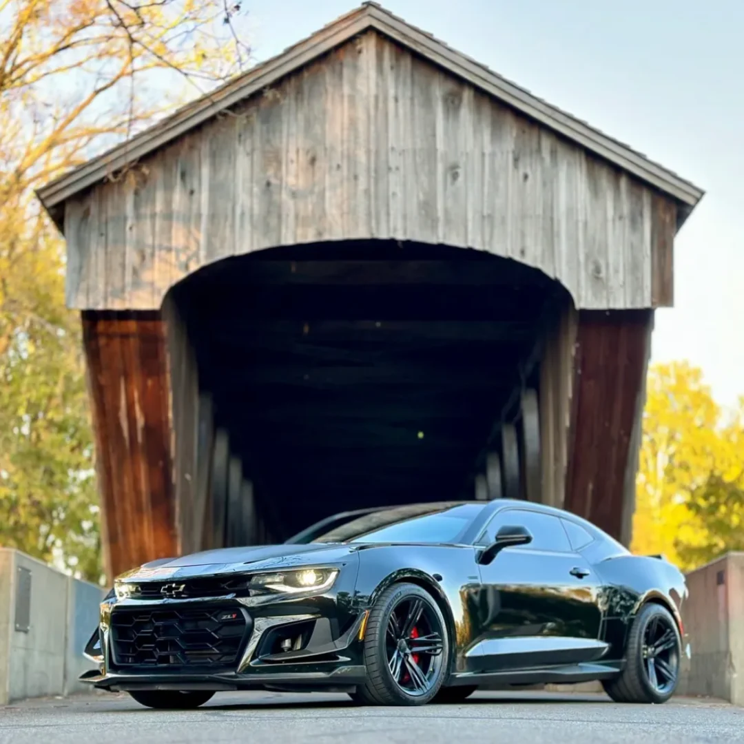 Sleek black sports car parked in front of a rustic wooden covered bridge.